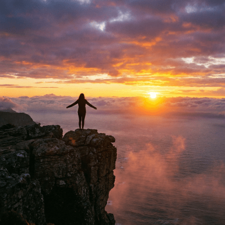 Silhouette of a person with outstretched arms standing on a cliff during sunset.